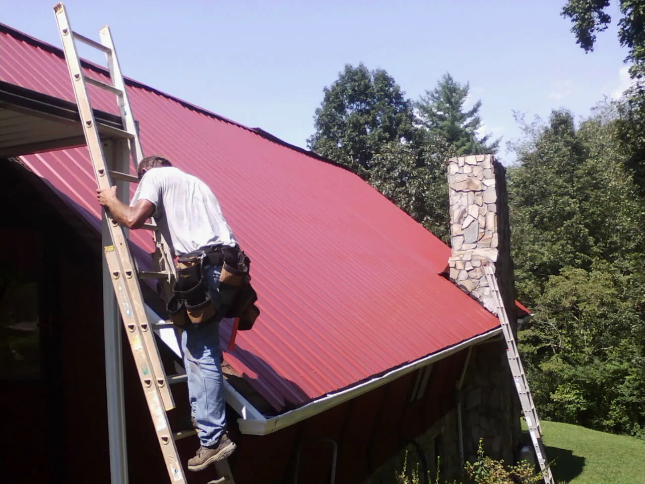 Roofer on a ladder beside a red metal roof and stone chimney — Soffit & Fascia Repair by Roofing Brothers in Yerington